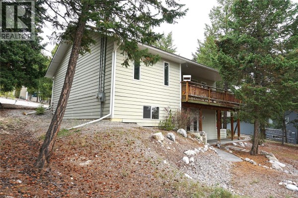 Side and back view of a light green house with a wooden deck and trees on a sloped lot. - 4969 Mountain Side Road, Fairmont Hot Springs