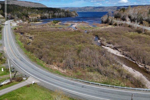 View of a curved paved road with a cleared lot and creek on the right, leading towards a large bay. - 49740 Cabot Trail, Goose Cove