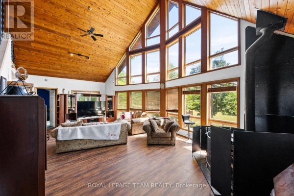 Living room with wood-paneled vaulted ceiling, large windows, dark wood floors, and comfortable furniture. - 4984 Dunrobin Road, Ottawa