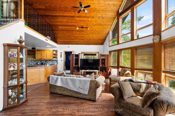 Interior view of a living room and kitchen area with wood accents, large windows, and a loft railing. - 4984 Dunrobin Road, Ottawa