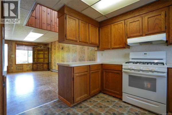 Kitchen corner with white gas range, wood cabinets, and view into dining area. - 4987 Gertrude St, Port Alberni