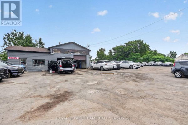 Grey commercial building with garage bays, parked cars, and a gravel lot. - 4997 & 4991 Bank Street, Ottawa