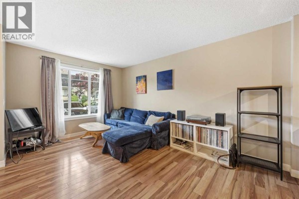 Living room with a blue sectional sofa, wood laminate floors, large window with grey curtains, and a television. - 5, 133 Copperpond Heights Se, Calgary