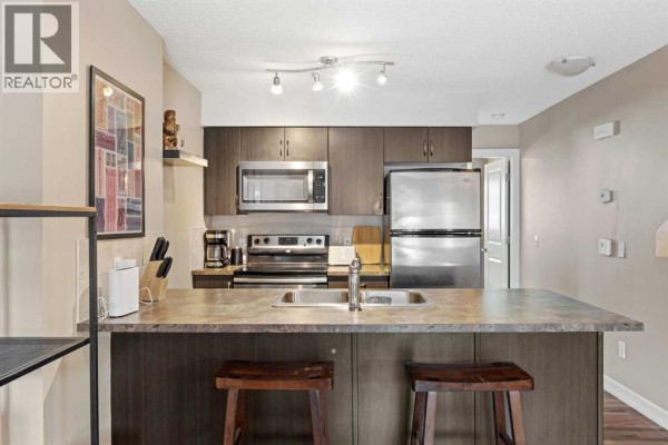 Kitchen with dark wood cabinets, stainless steel appliances, granite countertops, and a breakfast bar with stools. - 5, 133 Copperpond Heights Se, Calgary