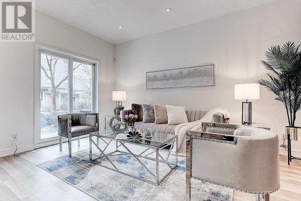 Living room with light wood floors, a grey sofa, silver armchairs, a glass coffee table, and large sliding glass door. - 5 - 21 St Moritz Way, Markham