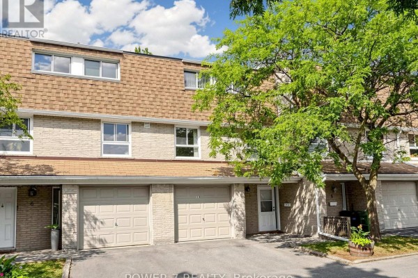 Close-up of townhouse exterior with garages, brick details, and white doors, surrounded by trees. - 5 - 77 Rameau Drive, Toronto