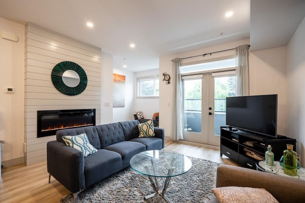 Living room featuring a dark blue tufted sofa, glass coffee table, electric fireplace, and television. - 5 46545 Brooks Avenue, Chilliwack Proper South, Chilliwack