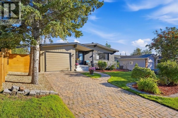 Exterior of a bungalow with a paved driveway, manicured lawn, and mature trees under a blue sky. - 5 Lake Huron Bay Se, Calgary
