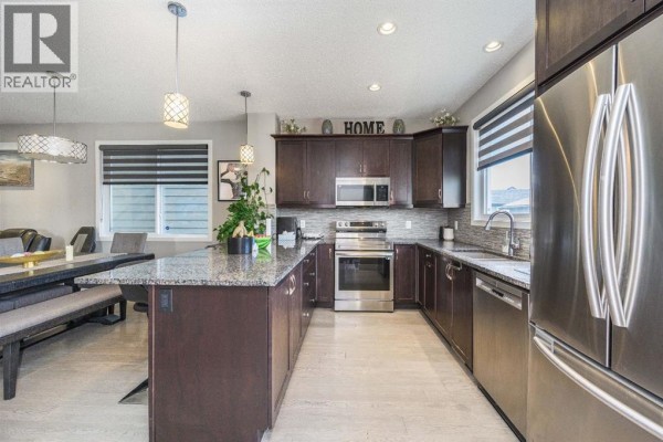 Open concept kitchen and dining area with dark brown cabinets, granite island, stainless steel appliances, and a dining table. - 5 Masters Street Se, Calgary