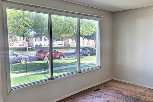 Living room with three large windows overlooking the street with parked cars and trees. - 50 Jerry Potts Boulevard W, Lethbridge