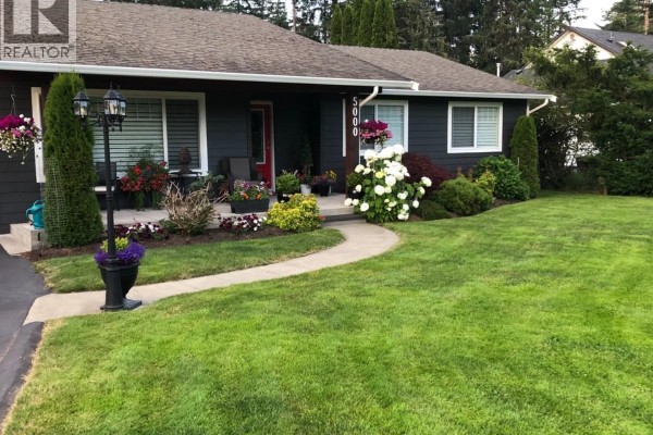 Front entrance of a dark grey house with white trim and a paved walkway with landscaping. - 5000 Childs Rd, Courtenay