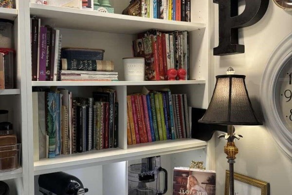 White built-in bookshelf filled with cookbooks, decorative items, and a black table lamp. - 5000 Childs Rd, Courtenay