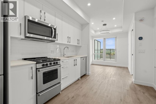Kitchen with white cabinets and stainless steel appliances, opening to a living area with large windows. - 502, 110 18a Street Nw, Calgary