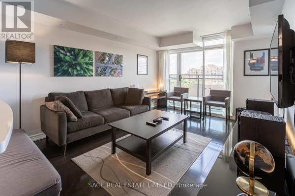 Living room with a dark grey sectional sofa, coffee table, chairs, and large window. - 502 - 120 Dalliomore Circle, Toronto