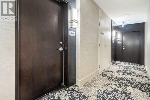 Apartment building hallway with dark wood doors, textured wallpaper, and patterned carpet. - 502 - 218 Queens Quay W, Toronto