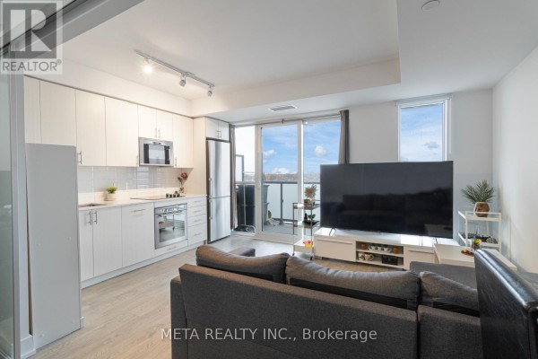 Apartment living area with a grey sectional sofa, entertainment unit, and kitchen visible. - 502 - 2550 Simcoe Street N, Oshawa