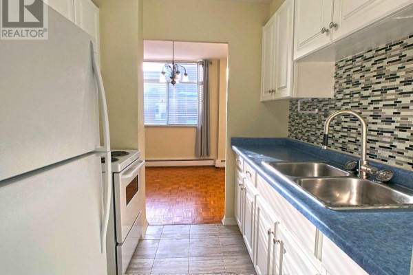 White kitchen cabinets, blue countertop, stainless steel sink, mosaic tile backsplash, and view of dining area with parquet flooring. - 502 - 335 Lonsdale Road, Toronto