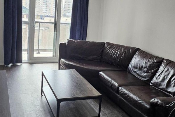 Living area with dark brown leather sectional sofa, coffee table, and dark curtains on a balcony door. - 502 - 8 Trent Avenue, Toronto