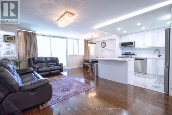 Living room with dark leather sofas, a patterned rug, and a view of a modern white kitchen. - 503 - 3590 Kaneff Crescent, Mississauga
