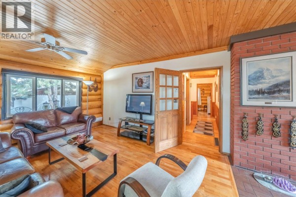 Living room with log walls, wood floors, a leather sofa, and a brick fireplace. - 5031 Gloinnzun Drive, 108 Mile Ranch