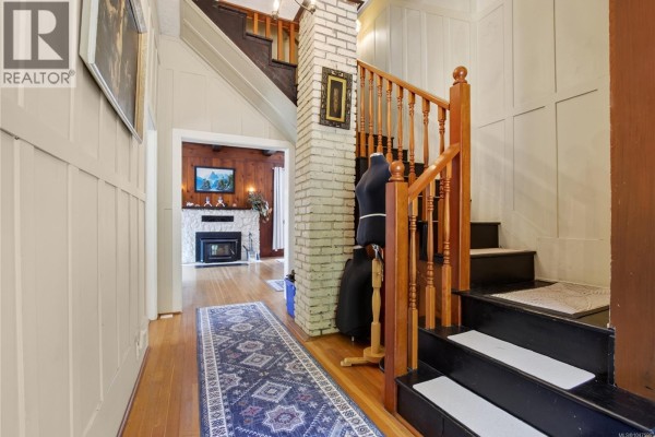 Hallway with wooden floors, stairs, brick fireplace, and doorways to other rooms. - 5034 Rocky Point Rd, Metchosin