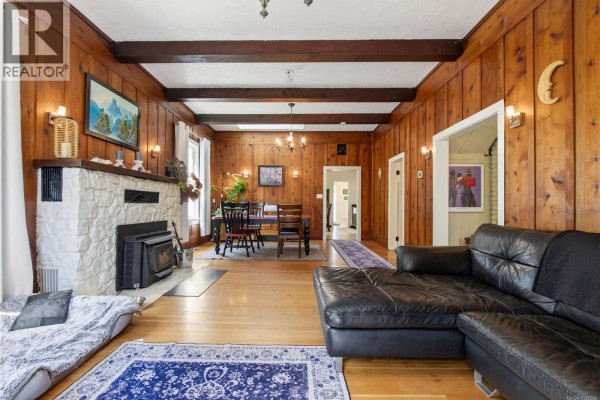 Living room with wood paneled walls, exposed beams, stone fireplace, and sectional sofa. - 5034 Rocky Point Rd, Metchosin