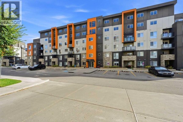 View of the apartment building's exterior and parking lot with cars under a blue sky. - 504, 10 Kincora Glen Park Nw, Calgary