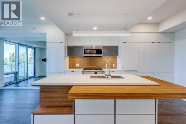 Kitchen island with a sink and wood countertop, facing stainless steel appliances and white cabinets. - 504, 1020 9 Avenue Se, Calgary