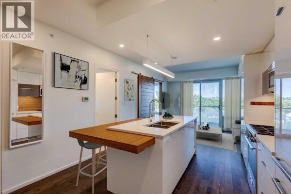 View of a kitchen island with seating, adjacent to a living area with a sofa and view of trees. - 504, 1020 9 Avenue Se, Calgary