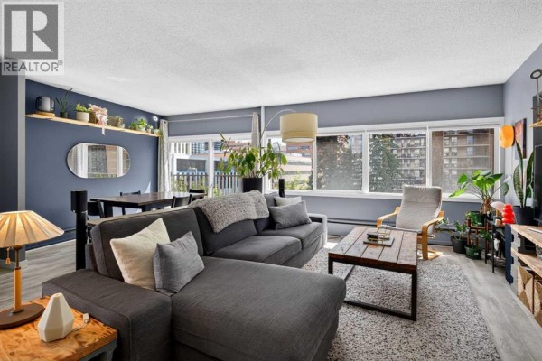 Living room with a gray sectional sofa, coffee table, large windows, and plants. - 504, 1209 6 Street Sw, Calgary
