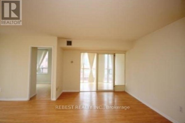 Living room showing light wood floors, cream walls, and a sliding glass door with sheer curtains leading outside. - 504 - 265 Enfield Place, Mississauga