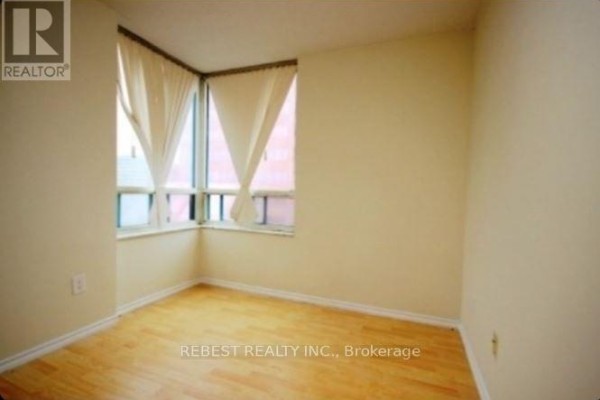 Empty bedroom with light wood floors, cream walls, and a large corner window with sheer curtains. - 504 - 265 Enfield Place, Mississauga