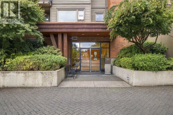 Close-up of the apartment building entrance with glass doors, brick accents, and surrounding greenery. - 504 1677 Lloyd Avenue, North Vancouver