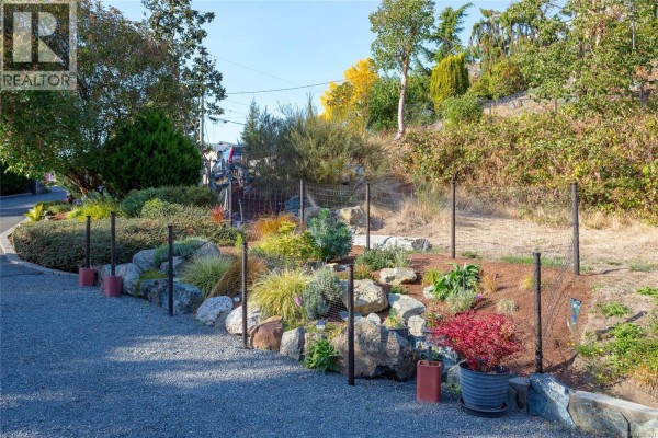Sloped garden bed with rocks, diverse plants, and black wire fencing along a gravel driveway. - 504 Langvista Dr, Langford