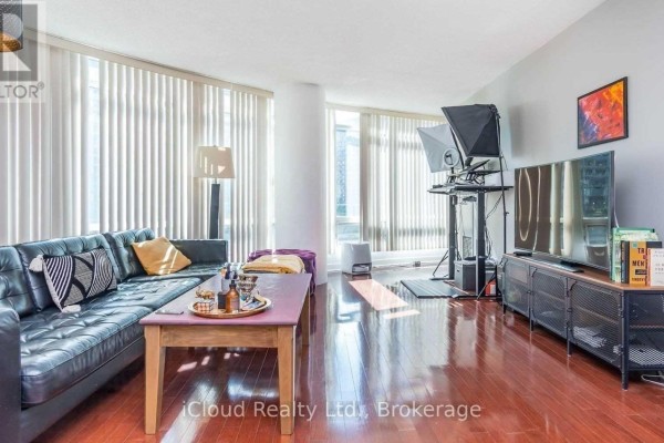Living room with a black leather sofa, wood floors, large windows with vertical blinds, and a television. - 505 - 373 Front Street W, Toronto
