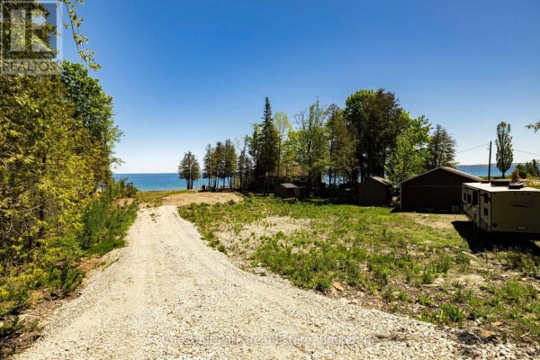 View from inside the property shows a gravel path, trees, an RV, and Georgian Bay in the distance. - 505107 Grey Road 1, Georgian Bluffs