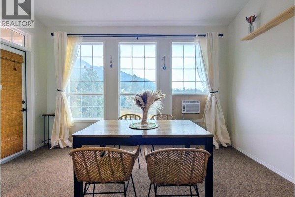 Dining area with large windows, mountain view, and a wooden table with rattan chairs. - 5052 Riverview Road Unit# 1013a, Fairmont Hot Springs