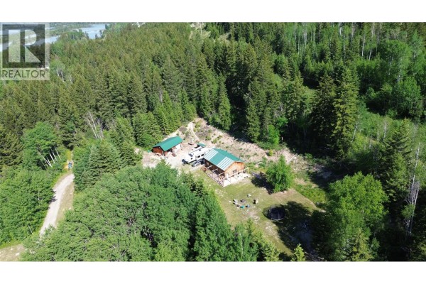 Grassy yard surrounded by tall trees, with a glimpse of wooden structures in the distance. - 5055 95 Highway, Castledale