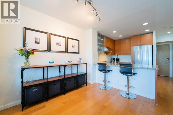 Open concept living area with a kitchen island, bar stools, framed artwork, and wooden floors. - 506 38 Front St, Nanaimo