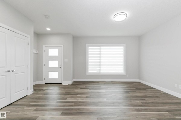 Empty living room with grey wood-look flooring, white walls, and a large window. - 5060,5062,5064 Chappelle Rd Sw, Edmonton