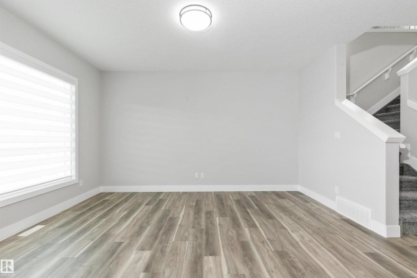 Empty room with grey wood-look flooring, white walls, and stairs leading up. - 5060,5062,5064 Chappelle Rd Sw, Edmonton