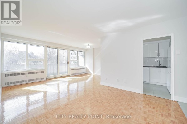 Living room with parquet floors and windows, view of the kitchen with white cabinets. - 507 - 10 Shallmar Boulevard, Toronto
