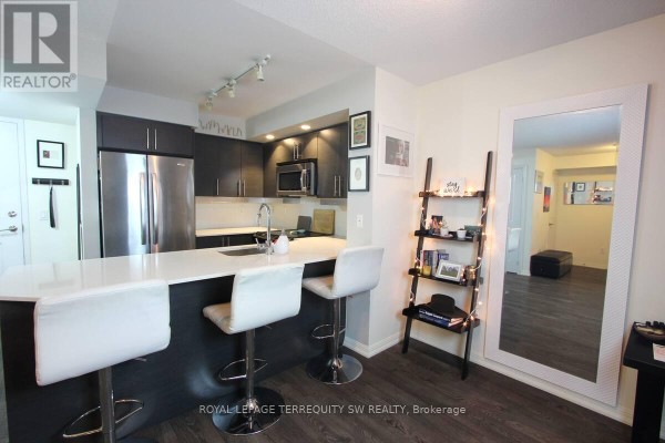 Kitchen island with white quartz countertop and three white stools, dark cabinetry. - 508 - 65 East Liberty Street, Toronto