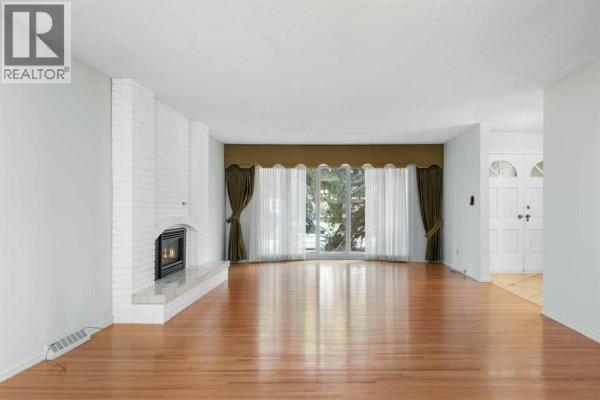 Living room featuring a white brick fireplace, hardwood floors, and large windows with drapes. - 508 Willow Brook Drive Se, Calgary