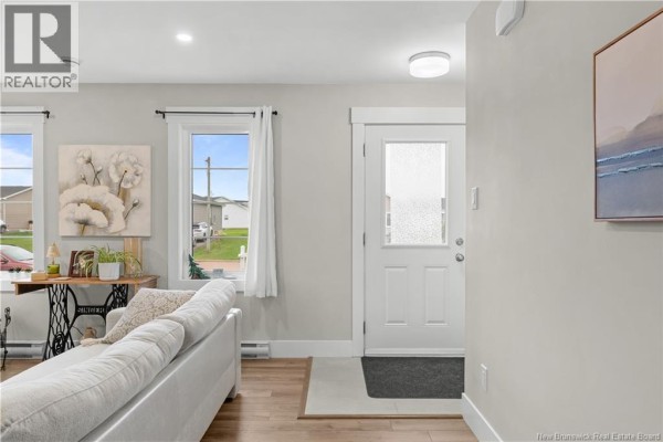 Living room with a white couch, wood floors, a window with white curtains, and a frosted glass front door. - 51-53 Macintosh Boulevard, Moncton
