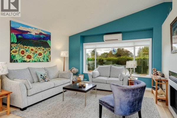 Living room with gray sofas, a dark coffee table, a fireplace, and a large window with a green view. - 51 901 Kentwood Lane, Saanich