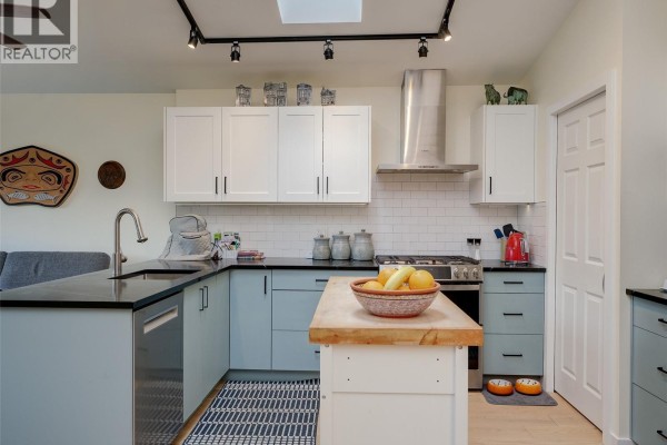 Kitchen with white and light blue cabinets, a stainless steel range hood, black countertops, and a central island. - 51 901 Kentwood Lane, Saanich