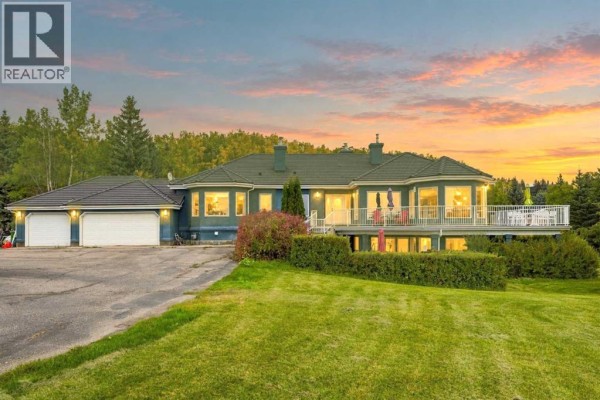 Blue house with a white deck and attached garage at sunset, with a lawn and trees in the foreground. - 51 Blueridge Close, Rural Rocky View County