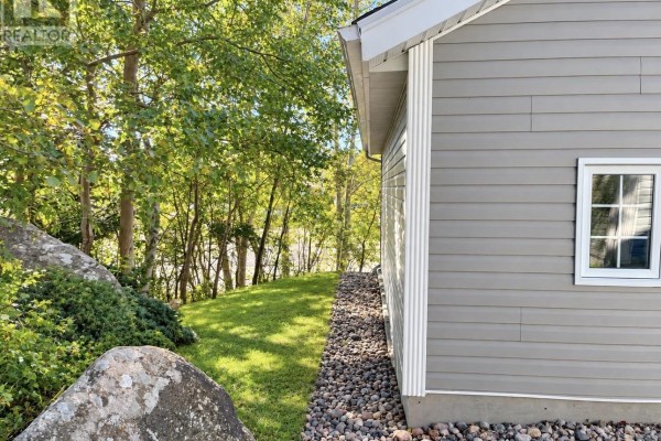 View of the side of the house and lawn leading towards a wooded area with large rocks. - 51 Main Street, Hare Bay