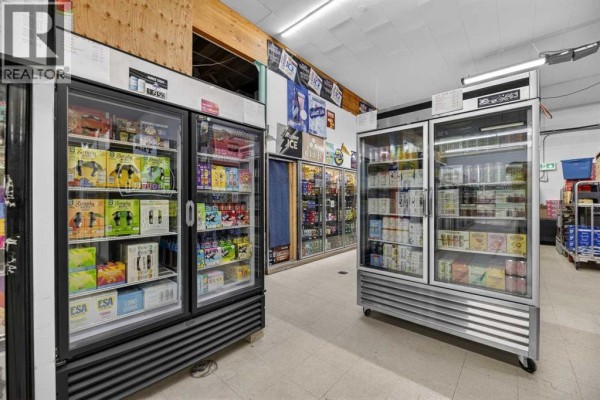 Interior view showing multiple glass-door refrigerators stocked with drinks, and shelving with various products. - 5107 50 Avenue, Olds